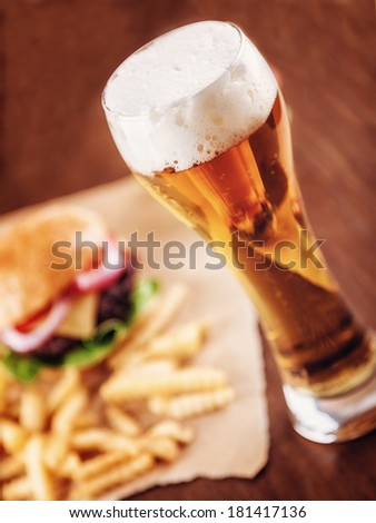 A glass of Beer with a Cheeseburger and Fries in the background. Focus is on Beer.