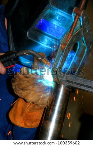 The TIG welder\'s protective helmet and leather glove are illuminated by the welding arc and sparks.