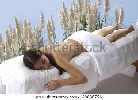 Pretty young brunette laying on massage table wrapped in a towel waiting on massage among a floral arrangement of tall ornamental grass in the background.