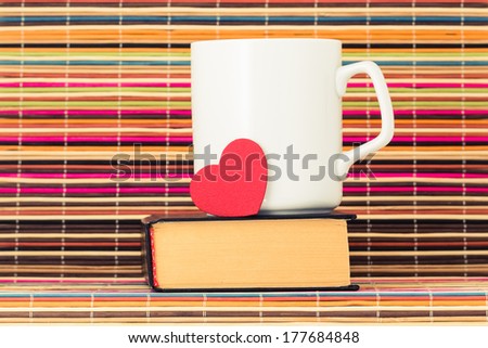 White cup with red heart stand on the book on a color background from bamboo sticks