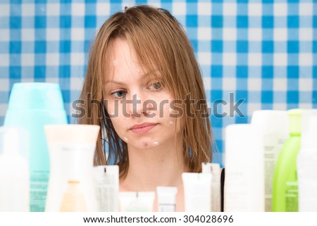 Young white-skinned girl with wet hair without makeup is looking at cosmetic products in bathroom. Skincare and beauty concept. Frontal portrait