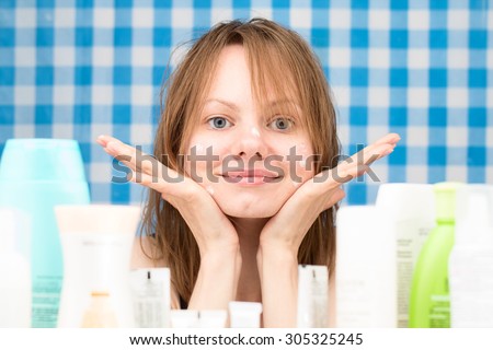 Young girl with wet hairs is posing in the bathroom in front of cosmetic products at blue-and-white checkered curtains background. Skincare and beauty concept. Frontal portrait