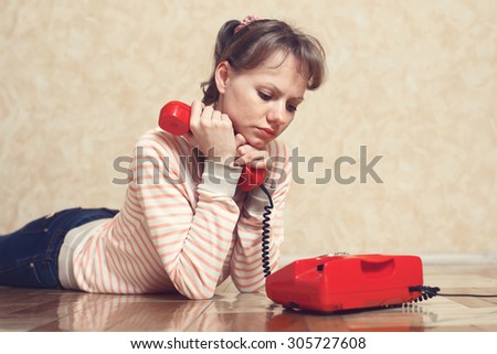Sad young white-skinned girl is sitting on parquet floor beside the wall near to red phone