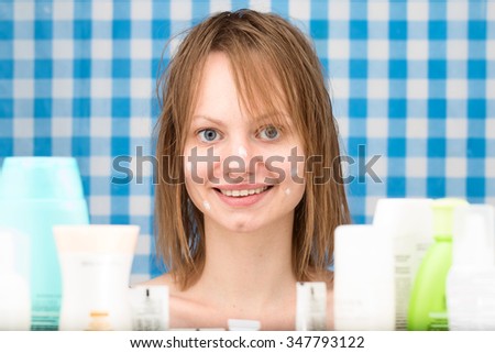 Girl without make-up with points of cosmetic product on her face is smiling surrounded by variety of cosmetics in the bathroom. Front portrait. Skin care and beauty concept