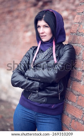 Outdoor fashion portrait of beautiful brunette woman posing in city street urban wall standing in the hood and a black leather jacket against a brick wall outdoors in spring