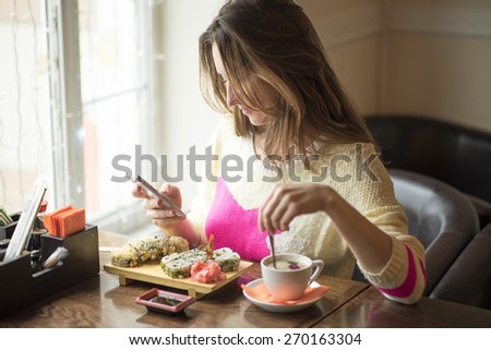 Young business woman eating sushi for lunch at a small caffe and a healthy lifestyle, working on a smartphone. Happy smiling, fashionable, in a yellow sweater.