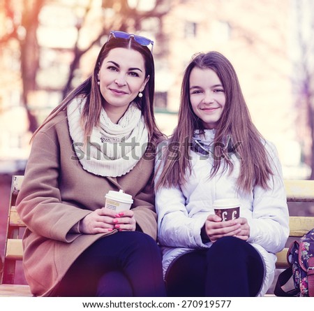 Mother and daughter sitting in autumn or spring park bench, are kept in the hands of cups of coffee or tea and smiling happy family.
