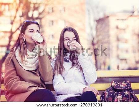 Mother and daughter sitting in autumn or spring in the park on a bench outdoors, drink coffee or tea or relax breakfast snack.