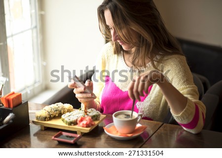 Young business woman eating sushi for lunch at a small cafe and a healthy lifestyle, working on a smartphone. Happy smiling, fashionable, in a yellow sweater.