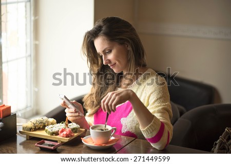 Young happy woman eating sushi for lunch at a small cafe and a healthy lifestyle, working on a smartphone. Smiling, fashionable, in a yellow sweater.