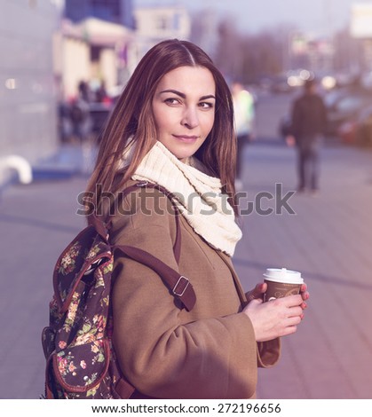 Portrait of a beautiful young brunette woman looking into the distance, in a coat and a scarf, standing in the street in spring or autumn. In his hand a cup of hot tea or coffee, fashion shoulder bag.