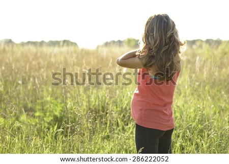 Pregnant girl standing in a field straightens hair, a bright sunny day a woman waiting for a child. In the meadow in a red dress with long hair.