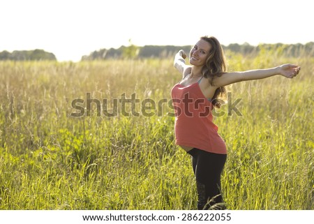 Young beautiful pregnant woman happy with open arms, in red a light summer dress, happy on the summer meadow in the grass in the bright sunny day. In anticipation of her child.