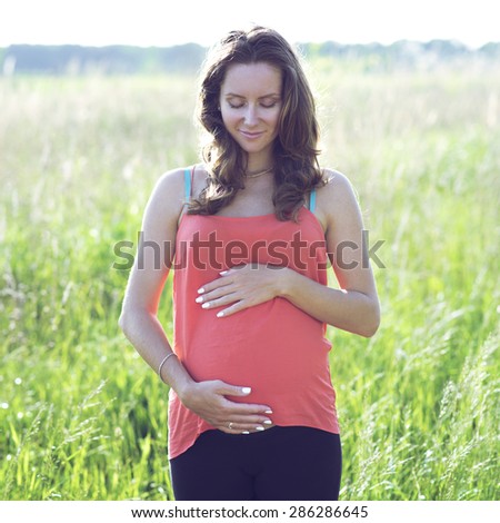 Happy beautiful pregnant woman holding belly and dreaming about the future of a small child, standing in a meadow on a bright sunny day in the green grass. Bright emotional.