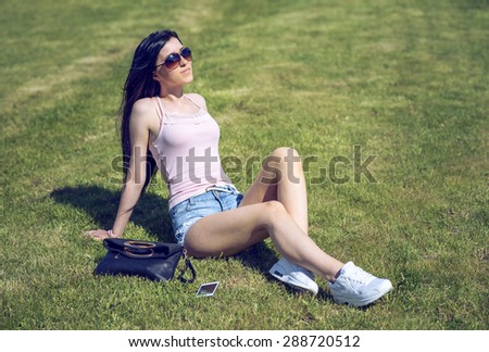 Beautiful female student relaxing in the fresh air on a bright sunny day, fashion style glamorous woman near black leather bag and a mobile phone, short denim shorts and sexy sun glasses happy resting