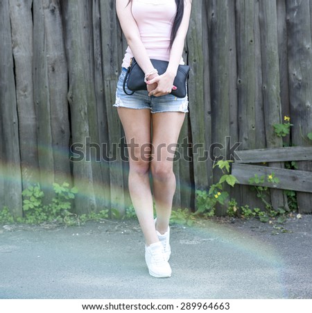 Fashionable girl holding bag posing in white sneakers, opposite the wooden fence with pink nail polish and bright glare from the sun