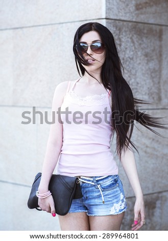 Sexy young brunette standing at the building, holding a black leather bag with pink nail polish, fashion glasses short denim shorts, rather slender