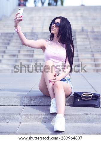 Brunette student photographs on your phone makes it self on the steps of the school institution in the open air on a bright sunny day, fashion style woman, a bag and a mobile phone, short denim shorts