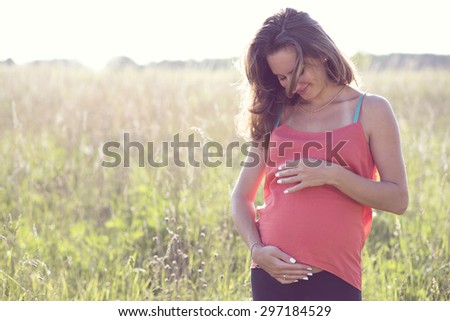 Pregnant brunette dreams in the bright sunny day thinking about the future of the child, smiling, holding belly with hands touching baby