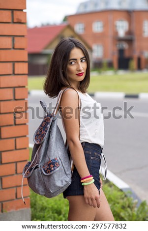 Young beautiful girl posing on brick wall background, with a white blouse and short jean shorts, fashion style bright summer on a street in the city is waiting with a bag
