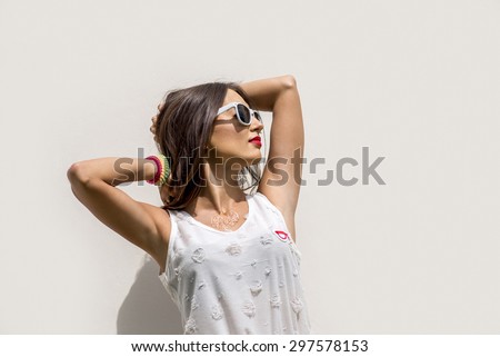 Bright summer fashion portrait of a young girl posing against a white wall, bright makeup brunette style glamor white sunglasses in the bright sun with a bracelet her arm with the tattoo on his chest