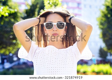 Beautiful young girl in glasses summer corrects hair, fashion style urban life. Brunette in the park bright summer day outdoors.