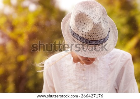 A young woman in retro-styled hat and lace blouse hiding face, trees in the background, sunset time