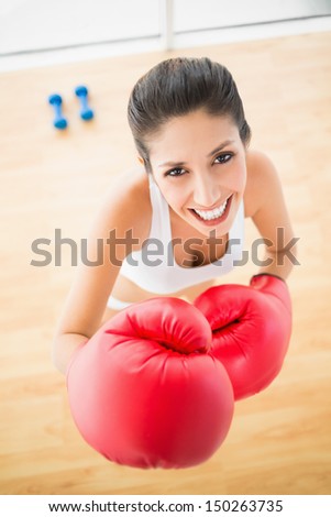 Fit woman wearing red boxing gloves smiling at camera at home in bright room