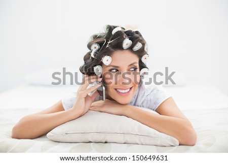 Happy brunette in hair rollers lying on her bed making a phone call in bedroom at home