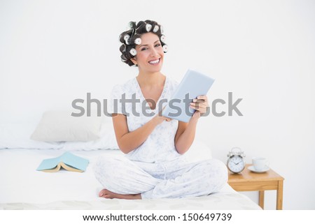 Pretty brunette in hair rollers using tablet on bed smiling at camera at home in bedroom