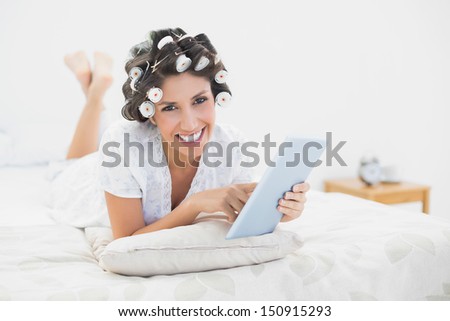 Smiling brunette in hair rollers lying on her bed using her tablet in bedroom at home