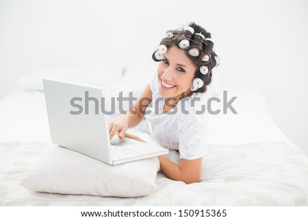 Happy brunette in hair rollers lying on her bed using her laptop in bedroom at home