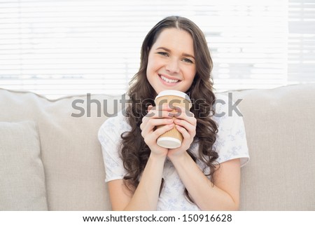 Cheerful pretty woman in pyjamas having coffee sitting on cosy sofa