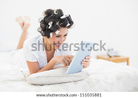 Cheerful brunette in hair rollers lying on her bed using her tablet in bedroom at home