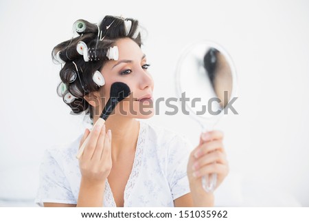 Pretty brunette in hair rollers holding hand mirror and applying makeup at home in bedroom