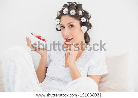 Sultry brunette in hair rollers having a bowl of strawberries at home in bedroom