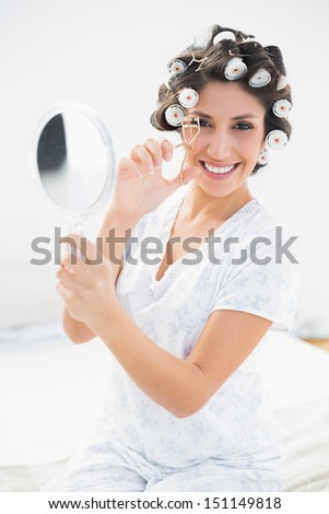 Pretty brunette in hair rollers holding hand mirror and using eyelash curler in bedroom at home