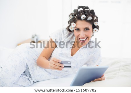 Happy brunette in hair rollers lying on her bed using her tablet to shop online in bedroom at home