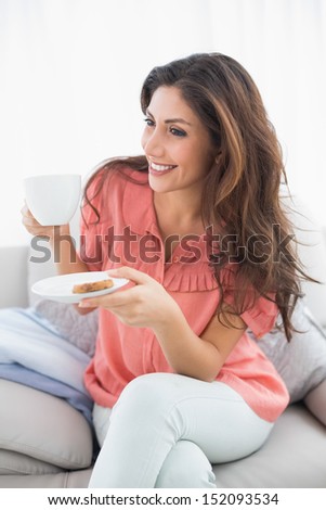 Pretty brunette sitting on her sofa holding cup and saucer at home in the sitting room
