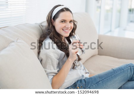 Gorgeous casual woman on cosy couch in bright living room holding glass of water