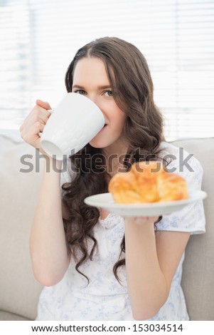 Cheerful young woman in pyjamas having breakfast while sitting on cosy sofa