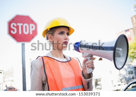 Frowning businesswoman wearing builders clothes holding megaphone outdoors on urban background