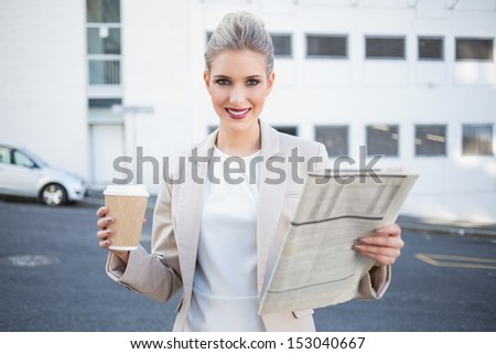 Smiling stylish businesswoman holding newspaper and coffee outdoors on urban background