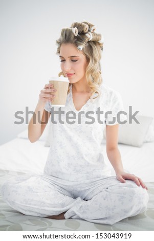 Peaceful pretty blonde wearing hair curlers smelling coffee sitting on cozy bed