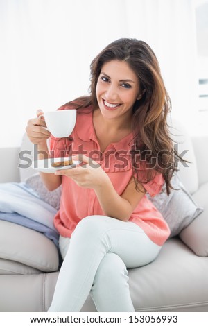Smiling brunette sitting on her sofa holding cup and saucer at home in the sitting room