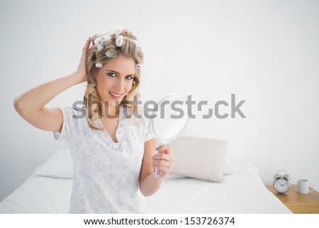 Cheerful gorgeous blonde wearing hair curlers holding mirror sitting on cosy bed