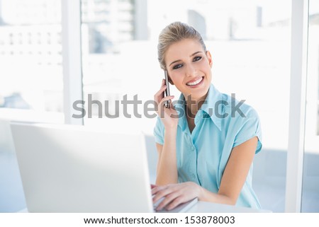 Happy smart businesswoman on the phone while using laptop in bright office