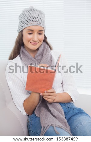 Happy pretty brunette with winter hat on writing on notebook sitting on cosy sofa
