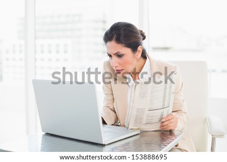 Stern businesswoman holding newspaper while working on laptop at the office