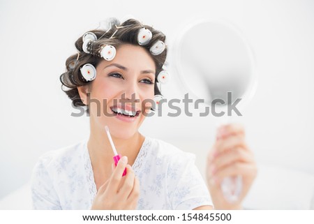 Happy brunette in hair rollers looking in hand mirror and holding  lip gloss at home in bedroom